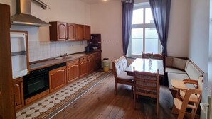 Une cuisine compacte avec des meubles en bois, un four encastré et un pare-feu en carreaux. On y trouve un coin repas avec une table et des chaises, ainsi qu’une fenêtre avec des rideaux.