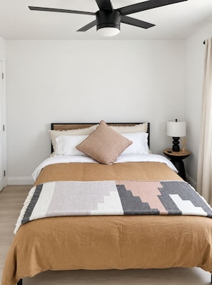 Un dormitorio con cama, ventilador de techo, mesita de noche con lámpara y ventana con cortinas.