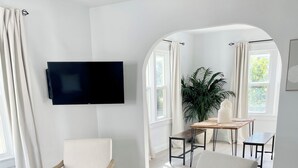 Un salon moderne avec un téléviseur à écran plat fixé au mur, deux fauteuils blancs, un pouf rond et un tapis à motifs.