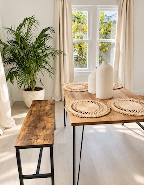 Un comedor con una mesa de madera, individuales tejidos, una planta en maceta y vistas de un jardín a través de una ventana.