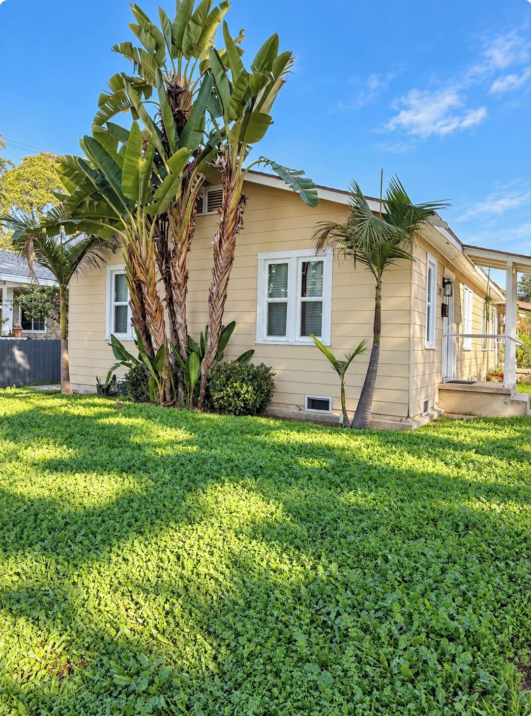 A single-story house with a front porch, a small garden with grass and a few plants, and a clear blue sky.