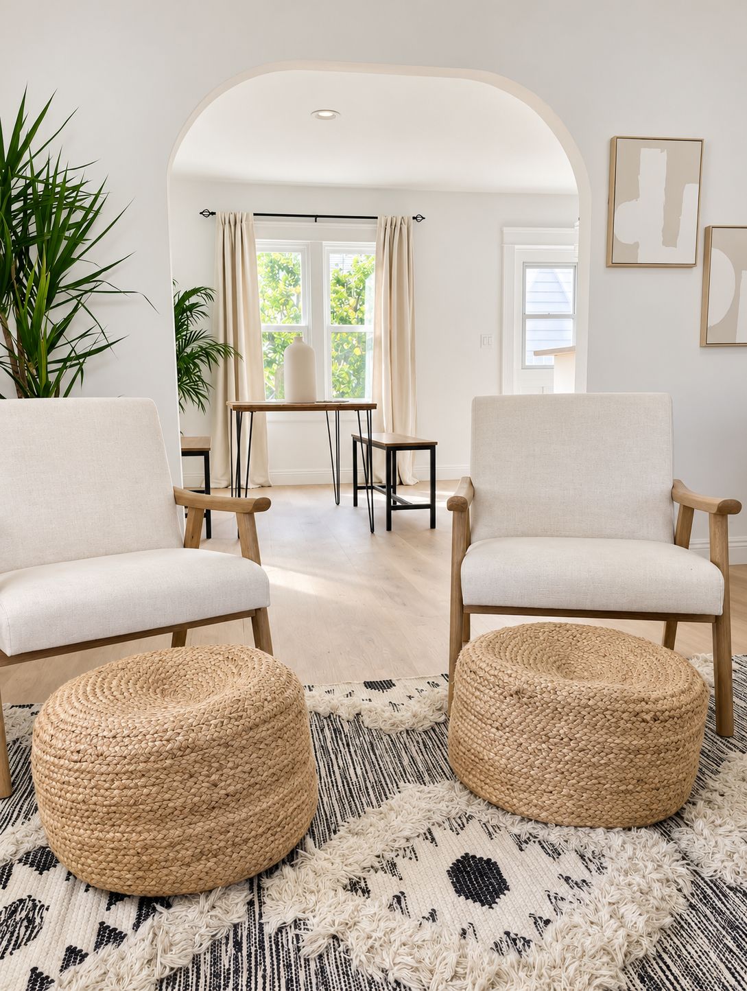A modern living room with two white armchairs, a round woven ottoman, a patterned rug, and a view through an arched doorway to a dining area with natural light.