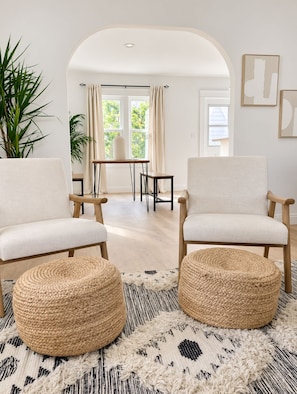 Una sala moderna con dos sillones blancos, un otoman tejido redondo, un tapete estampado y vistas a un comedor con luz natural a través de un arco.