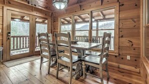Interior de cabana de madeira com mesa de jantar e cadeiras, grandes janelas com vista para árvores e montanhas, e piso de madeira.