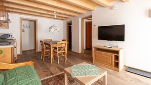 A cozy living room with a wooden ceiling, a dining area with wooden chairs, a television on a stand, and a microwave on a cabinet.