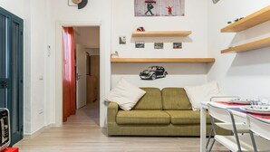 Uma sala de estar com um sofá verde, uma mesa de jantar com cadeiras, estantes de madeira e um relógio de parede.