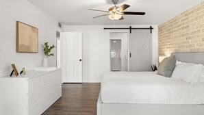 Ein Schlafzimmer mit Bett, Deckenventilator, weißem Schminktisch, Pflanze und Backsteinwand.
