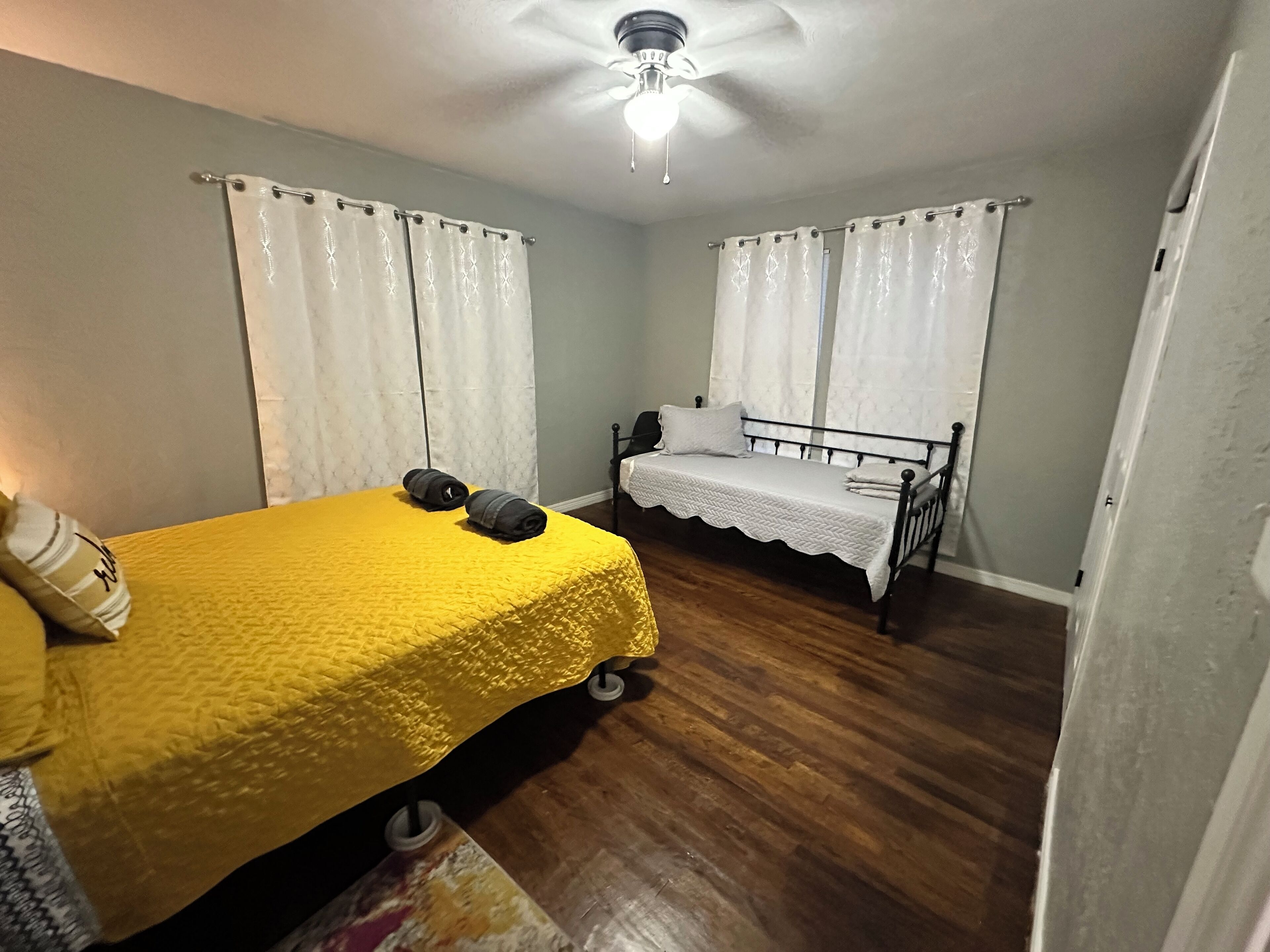 A bedroom with a yellow bedspread, two rolled towels, a white bed with a white bedspread, and a ceiling fan.