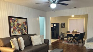 A living room with a brown leather sofa, a gray ottoman, and a colorful rug. There is a ceiling fan, a dining area with a table and chairs, and a refrigerator.