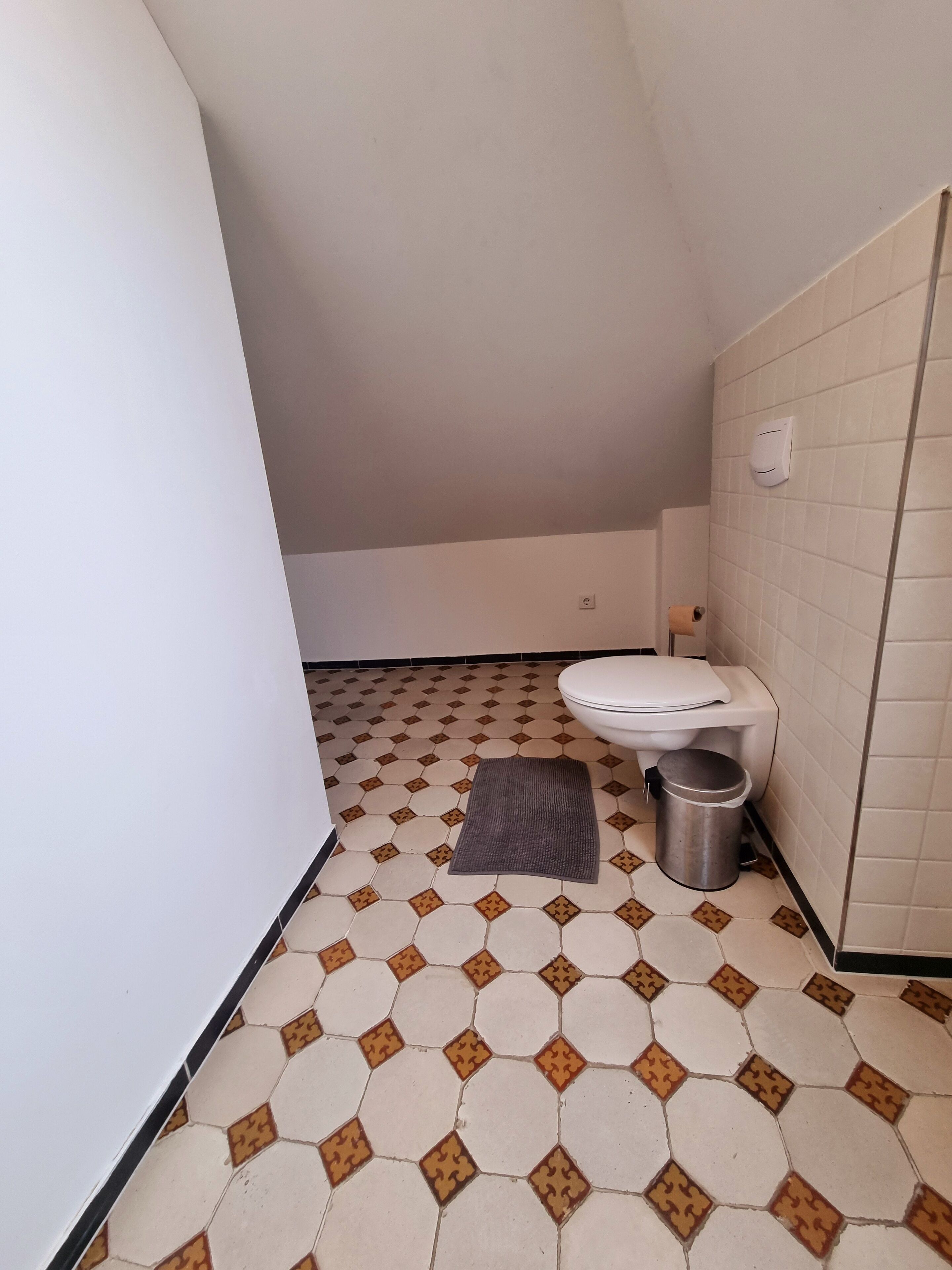 A small, narrow bathroom with a toilet, a trash bin, and a patterned floor.
