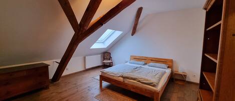 Un dormitorio con techo inclinado, tragaluz, cama de madera, una silla y un radiador.