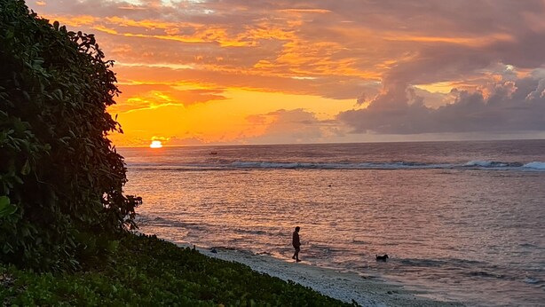 Een zonsondergang boven de oceaan, met een persoon die langs het strand loopt.