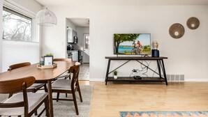 A dining area with a wooden table and chairs, a pendant light, a painting of cyclists, and a small plant on a console table. - Bay Breeze Getaway | Blocks to the Beach | Downtown | AC | Fenced Yard (Traverse City)