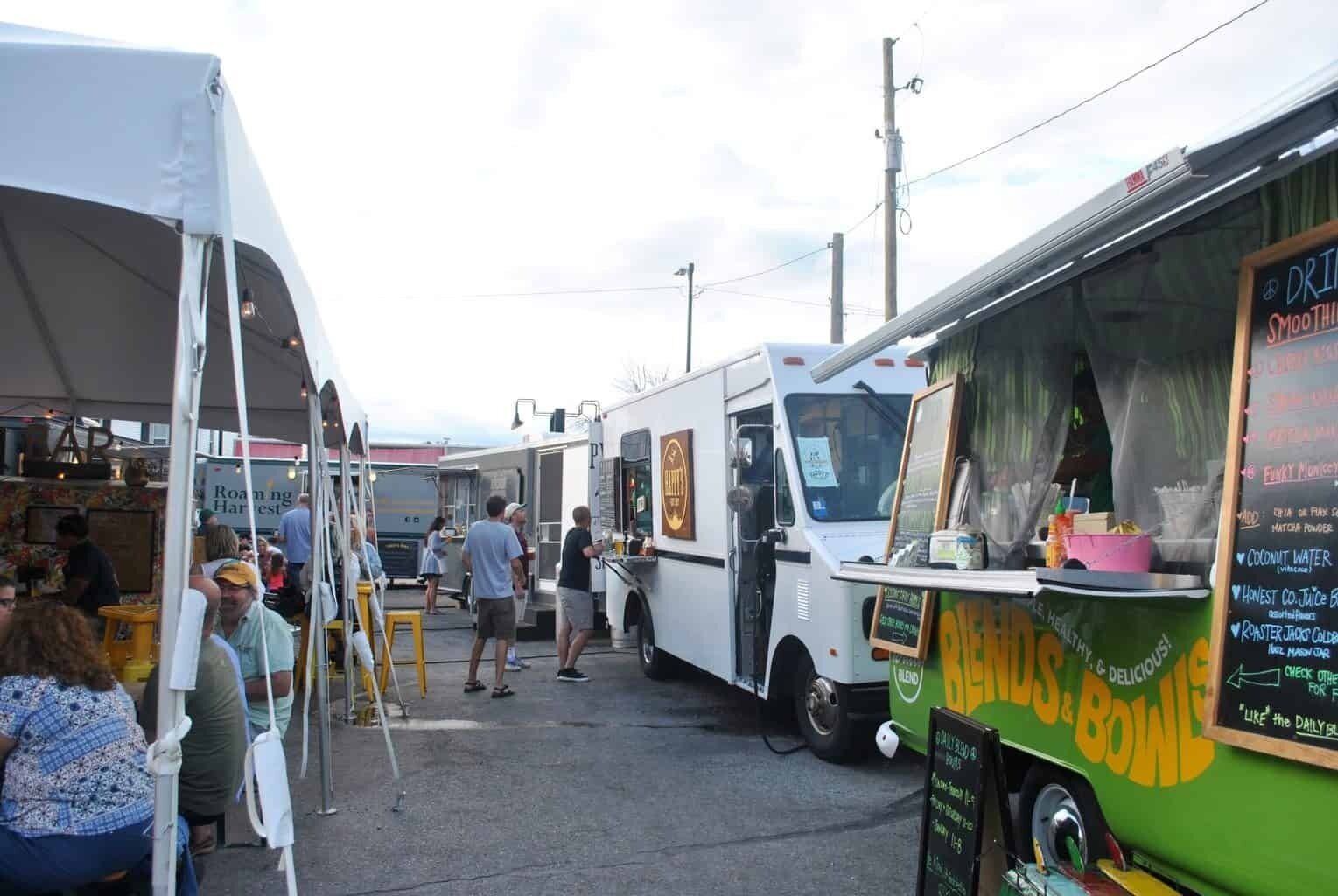 A food truck market with multiple food trucks parked in a row, people walking around, and a tent in the background.
