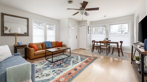A living room with a sofa, coffee table, rug, and a dining area with chairs and a table. - Bay Breeze Getaway | Blocks to the Beach | Downtown | AC | Fenced Yard (Traverse City)
