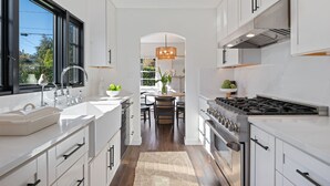 A modern kitchen with white cabinetry, a central island, a large window, and a view of the outdoors. - Charming Urban Bungalow with Garden and Deck (Santa Barbara)