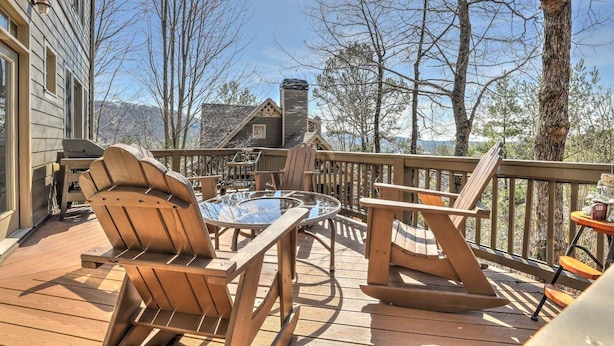 En träveranda med Adirondack-stolar, ett bord med glasskiva och en eldstad i sten.
