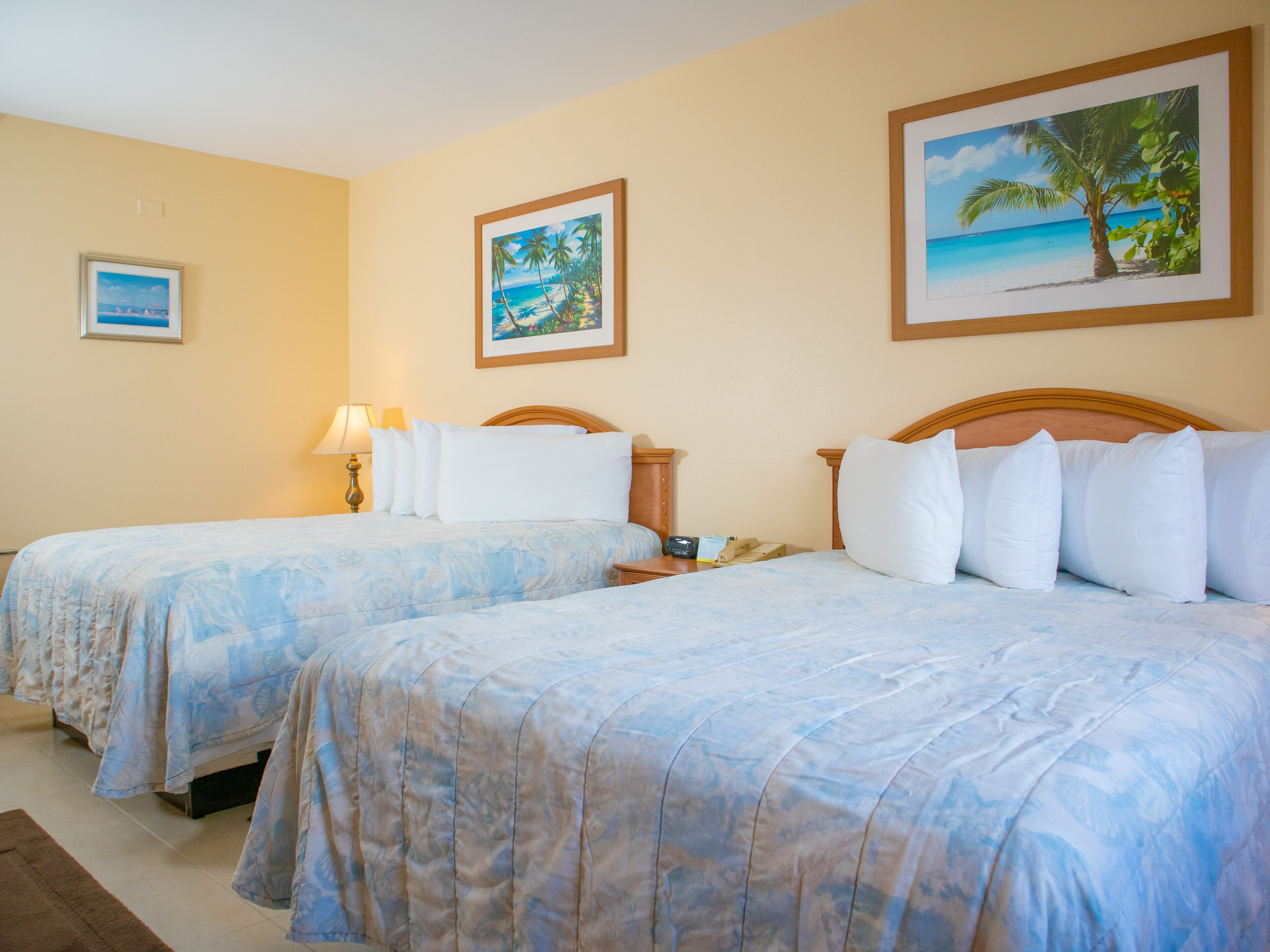 Habitación de hotel con dos camas, paredes color beige y cuadros enmarcados con escenas tropicales.