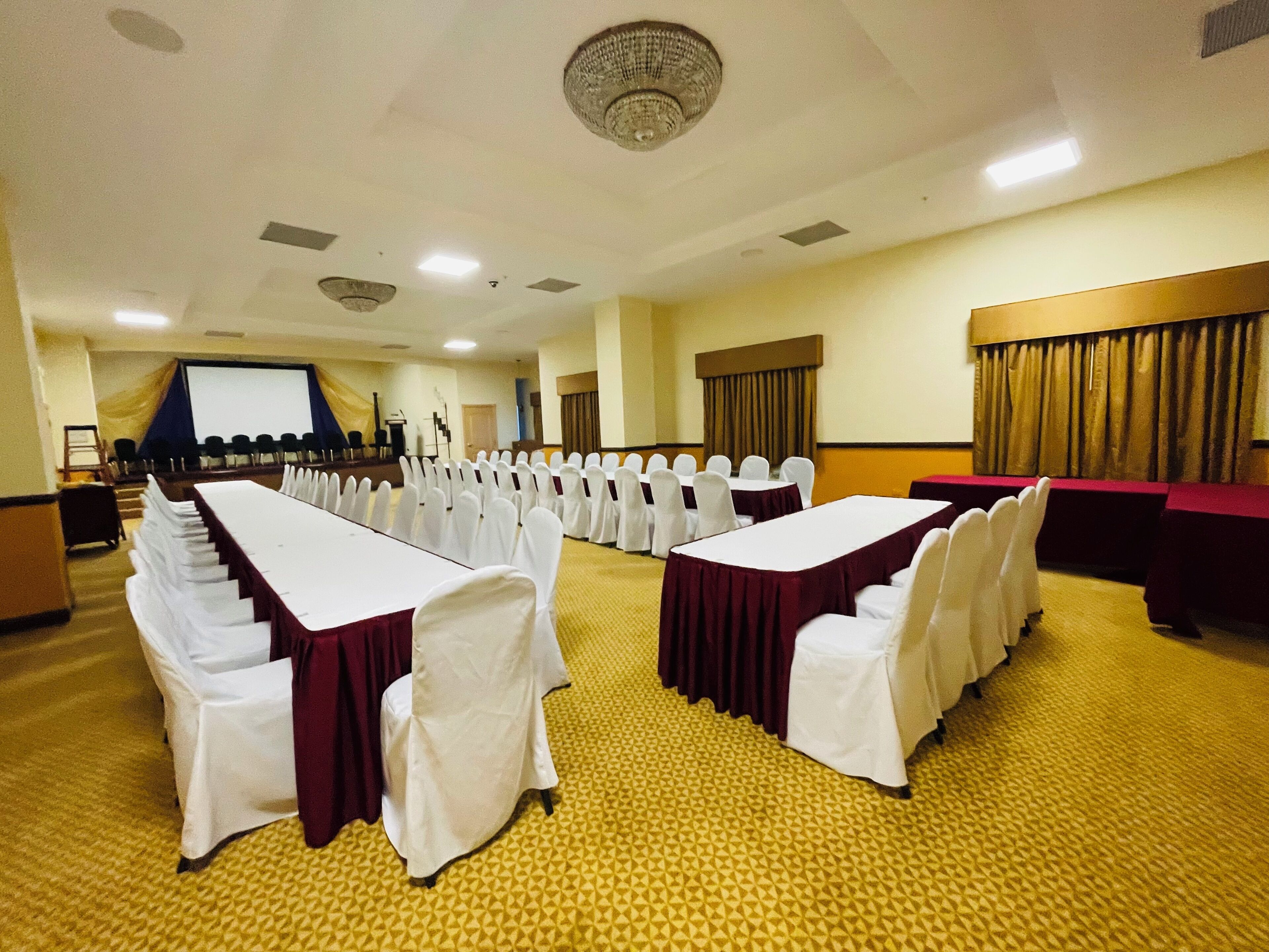 A conference room with long tables and chairs, a stage with a screen, and a carpeted floor.