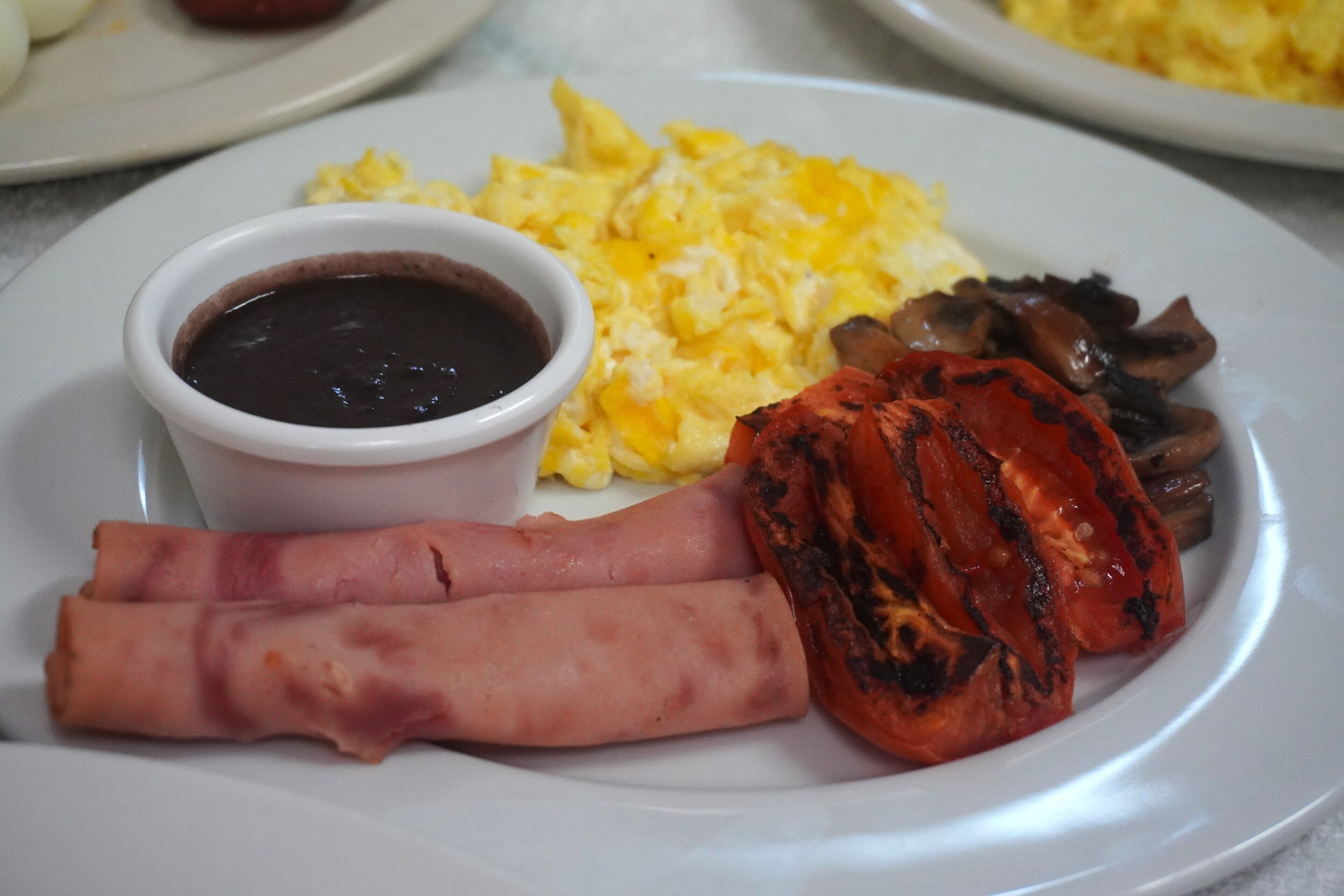 A plate with scrambled eggs, grilled tomatoes, a slice of ham, and a small bowl of sauce.