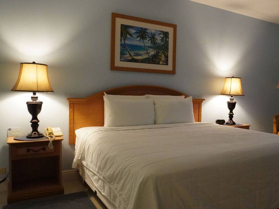 Un lit bien fait, avec du linge de lit blanc, deux lampes de chevet, une table de chevet en bois avec un téléphone, et un tableau représentant une scène de plage accroché au mur.