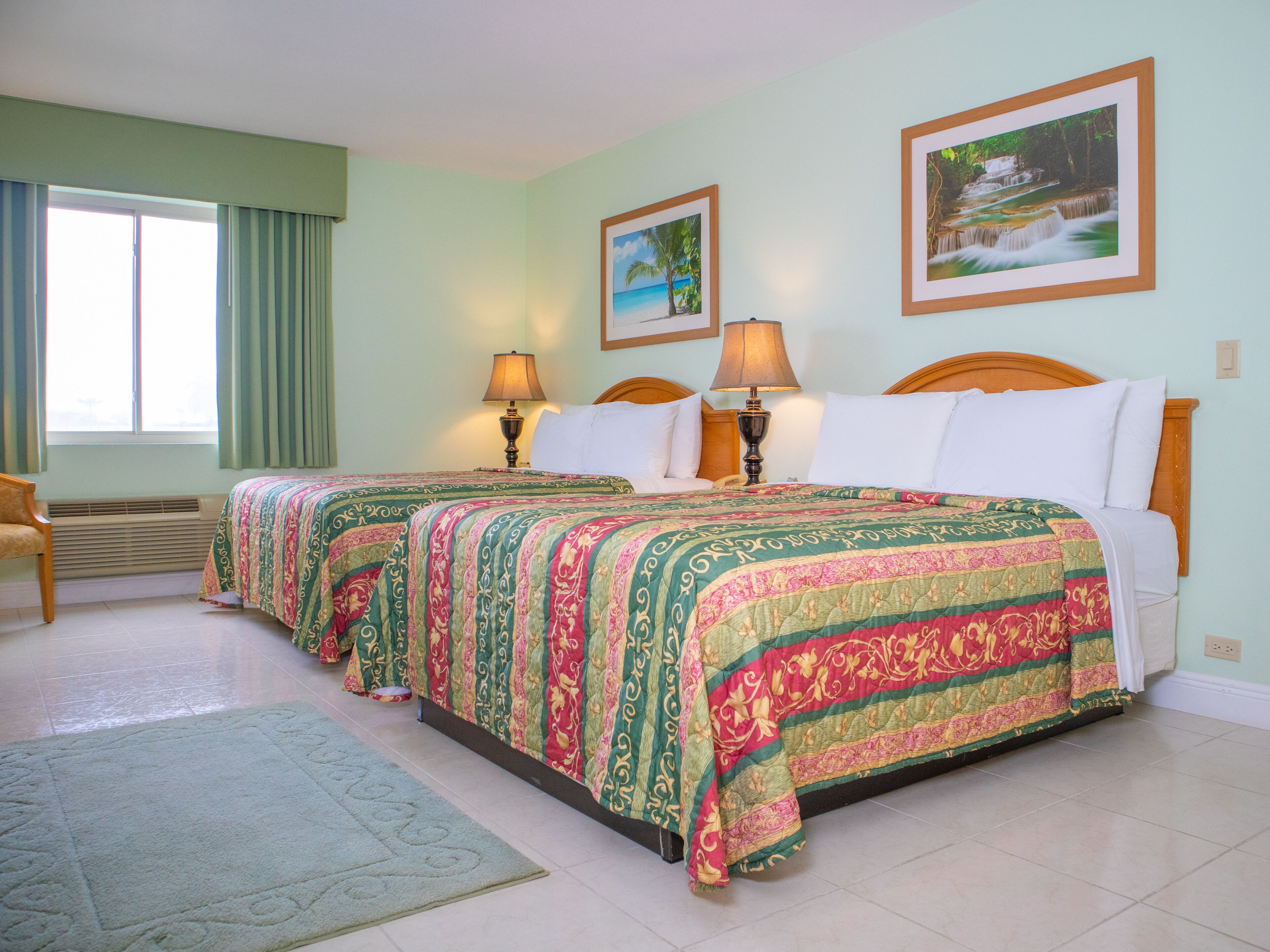A hotel room with two beds, a window with curtains, a radiator, and two framed pictures on the wall.