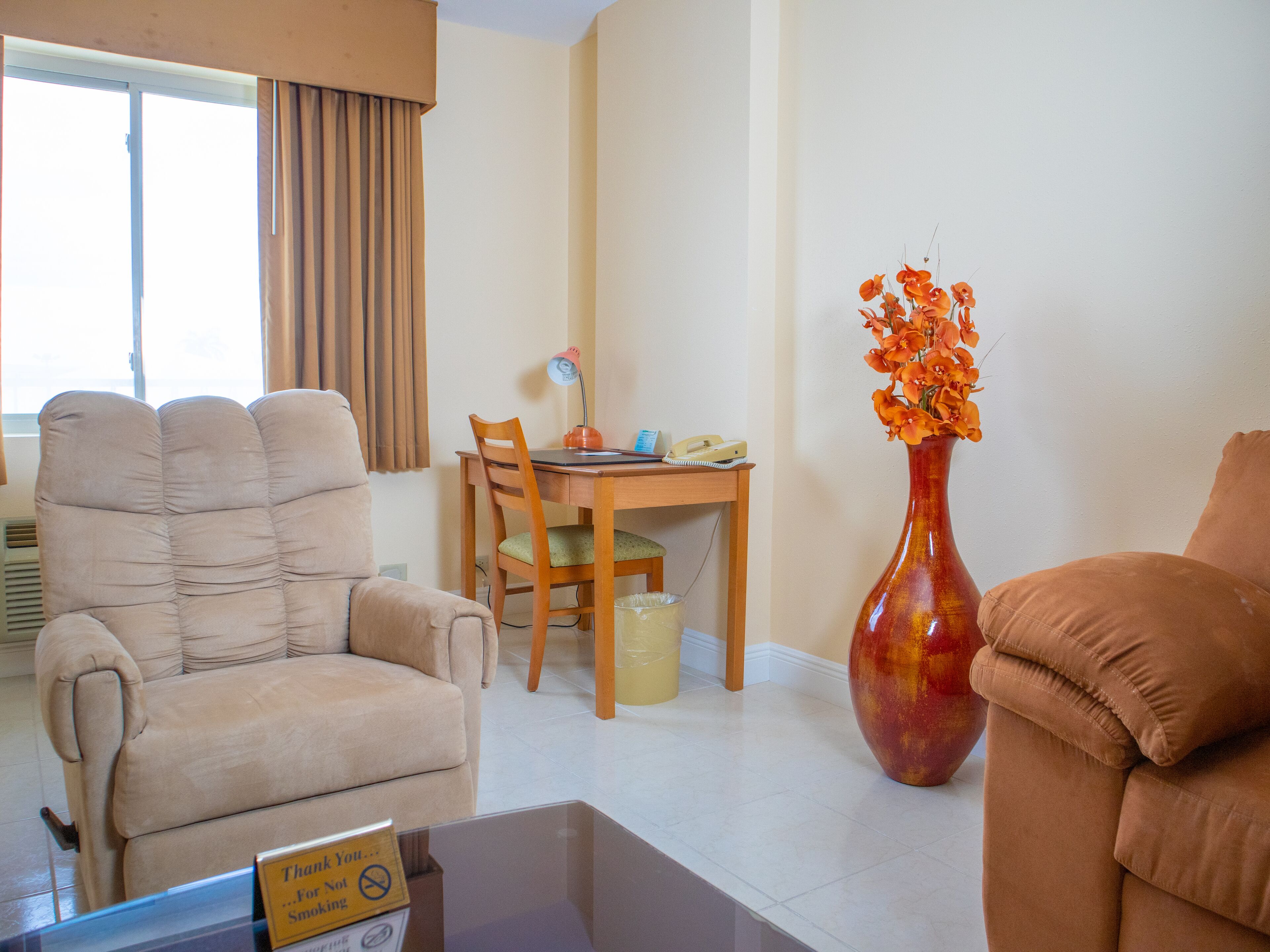 A living room with a beige armchair, a wooden dining table with a chair, a vase with orange flowers, and a sign indicating no smoking.