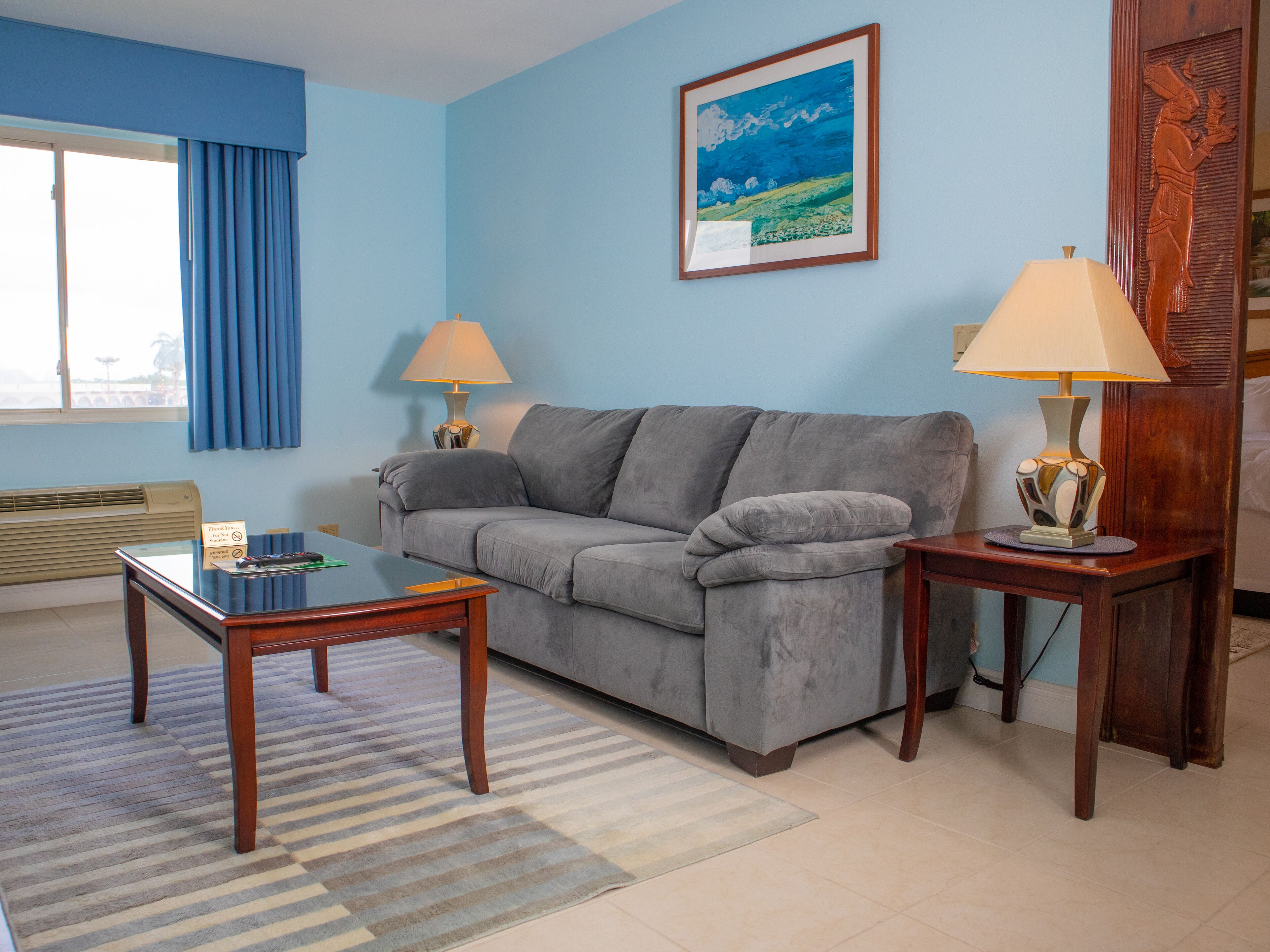A living room with a grey sofa, two side tables with lamps, a framed painting, and a window with blue curtains.