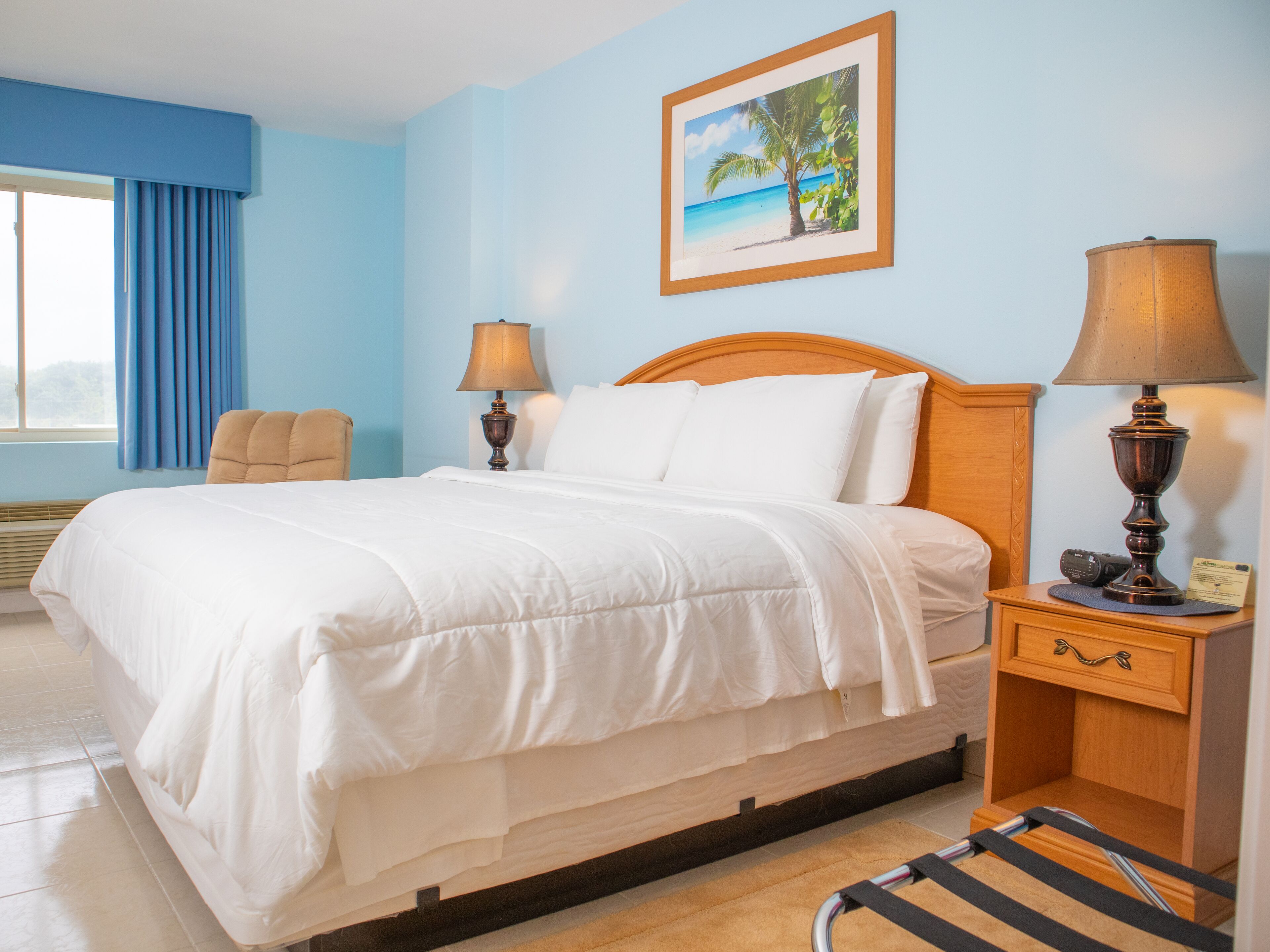 A hotel room with a bed, two bedside lamps, a nightstand, a chair, a window with curtains, and a painting of a beach scene.