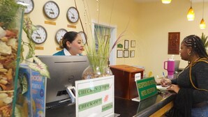A hotel reception desk with clocks displaying different time zones, a check-in/out sign, and a desk with a computer and a plant. - Princess Vegas Hotel & Casino (Corozal)