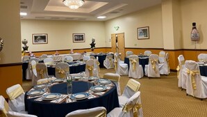 A banquet hall with round tables set for a formal event, featuring white chairs with gold sashes, blue tablecloths, and a chandelier. - Princess Vegas Hotel & Casino (Corozal)