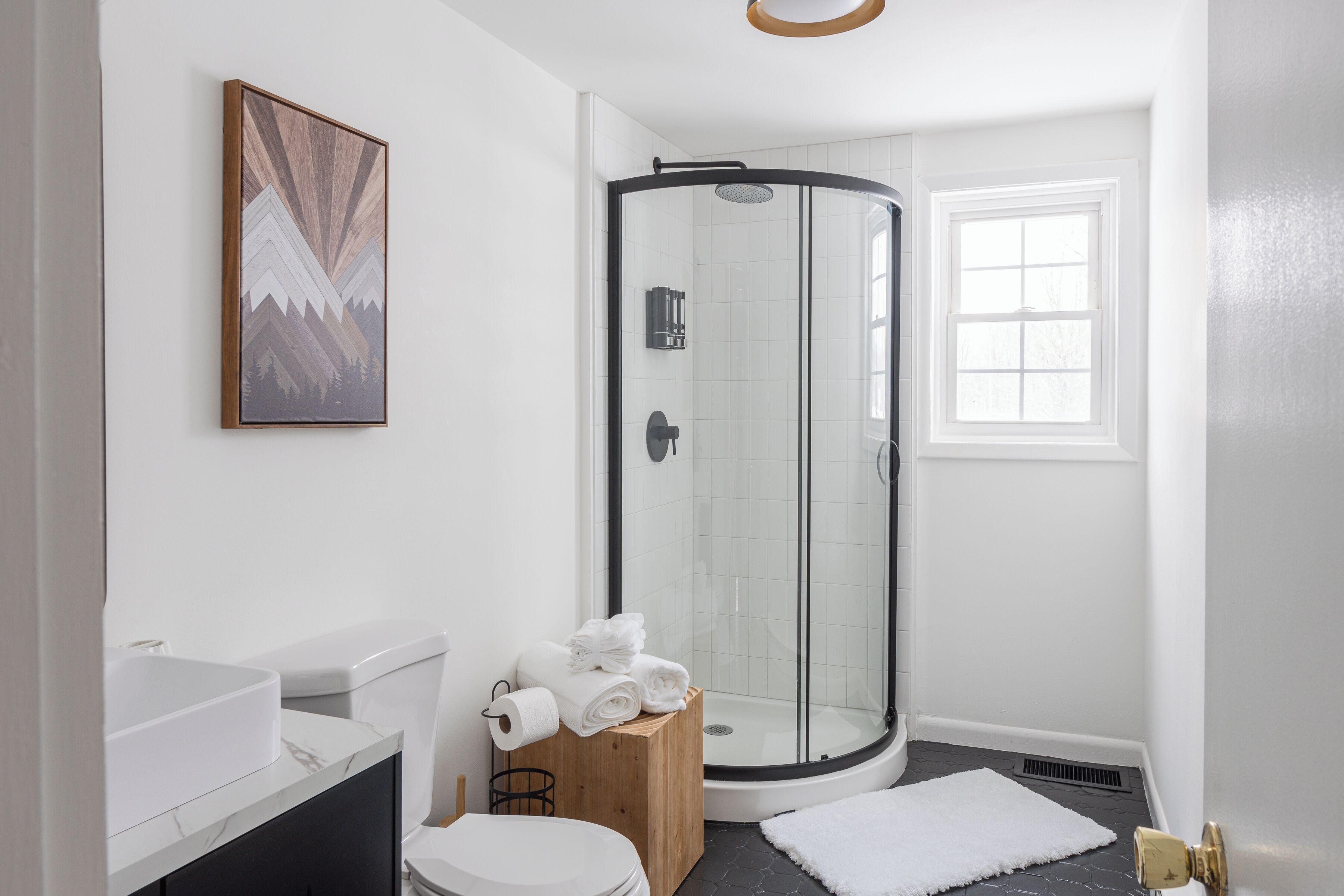 Een moderne badkamer met een douchecabine van glas, een witte wastafel en een houten kruk met handdoeken.