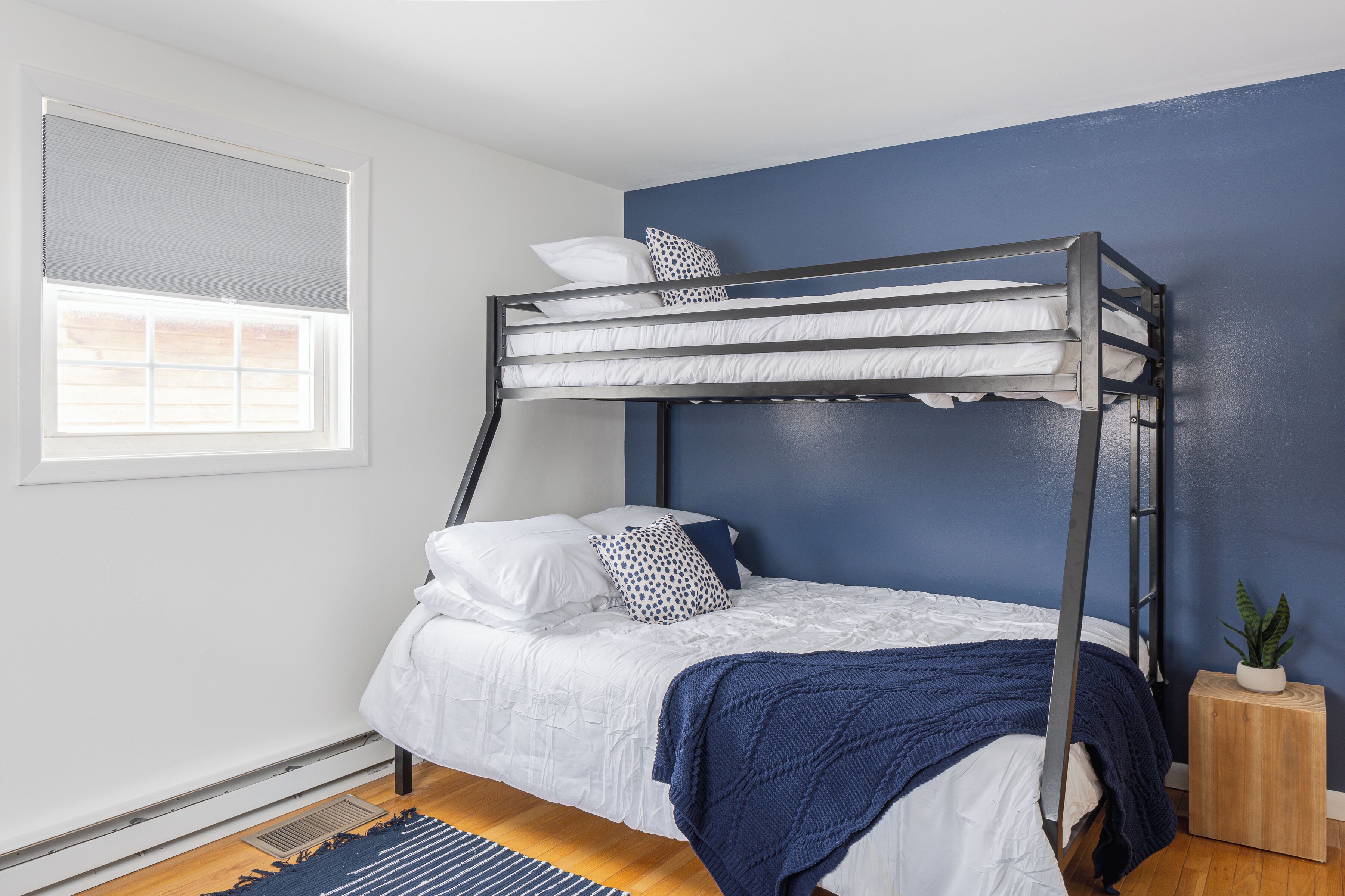Una cama litera con ropa de cama blanca y una manta azul, una mesita de madera con una planta, y una ventana con persianas.