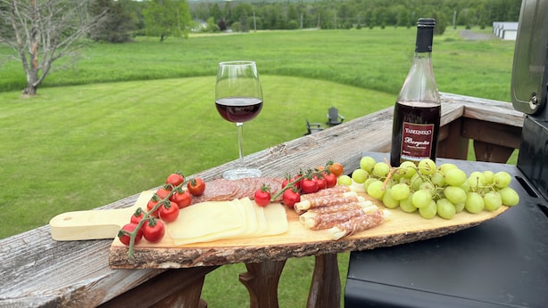 A picnic setup with a wine glass, bottle, cheese, tomatoes, and grapes on a wooden table with a view of a grassy field and hills.
