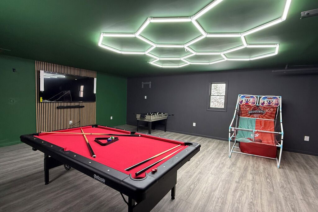 Une table de billard recouverte d’un drap de feutre rouge, une table de baby-foot et un jeu d’arcade de basketball dans une pièce aux murs verts et dotée d’un luminaire hexagonal au plafond.