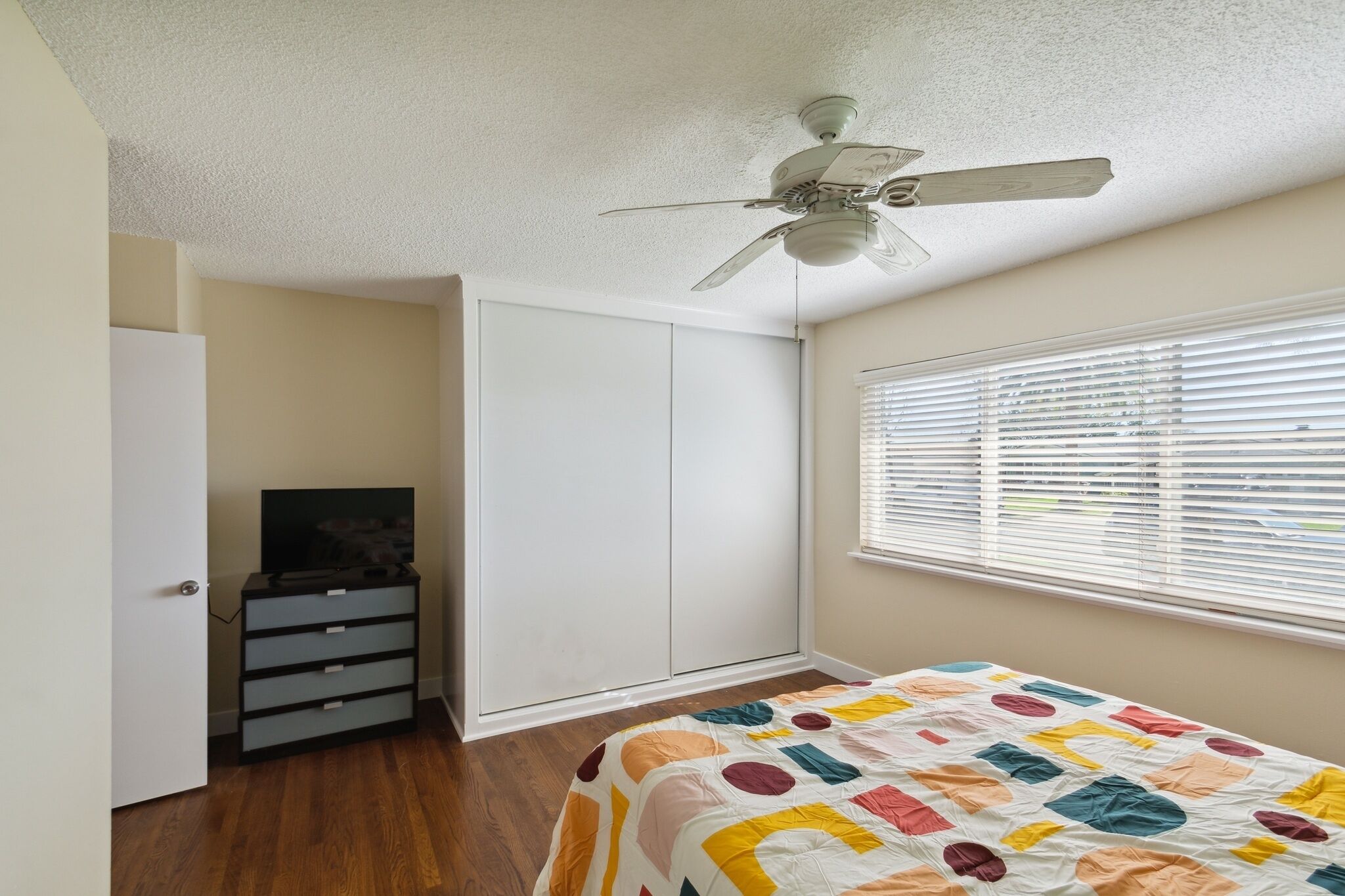 A bedroom with a bed, a ceiling fan, a wardrobe, a dresser, and a window with blinds.