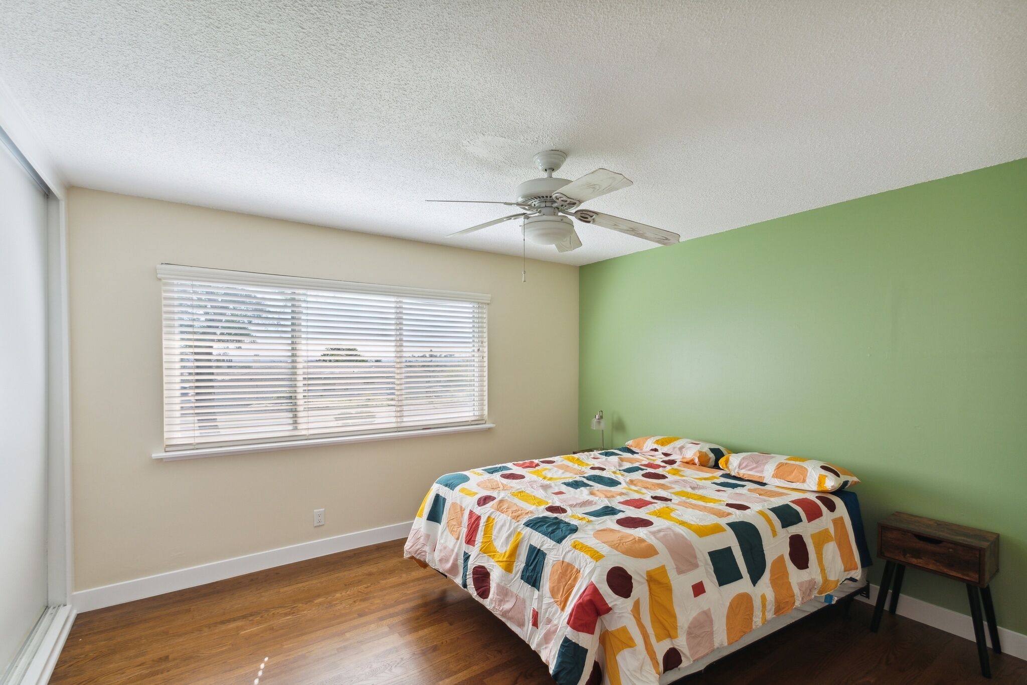 Une chambre avec un lit, un ventilateur de plafond, une fenêtre avec des stores et un mur vert.