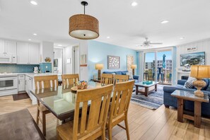 A modern kitchen with white cabinets, a dining area with wooden chairs and a glass table, a living room with a blue sofa and a television, and a balcony with a view of buildings.