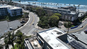 Um edifício alto com área de terraço no telhado, uma estrada curva com faixa de ônibus e uma praia com um píer ao fundo.