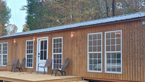 Une cabane en bois dotée de grandes fenêtres, d’une terrasse avec des chaises et d’une lanterne sur le toit.