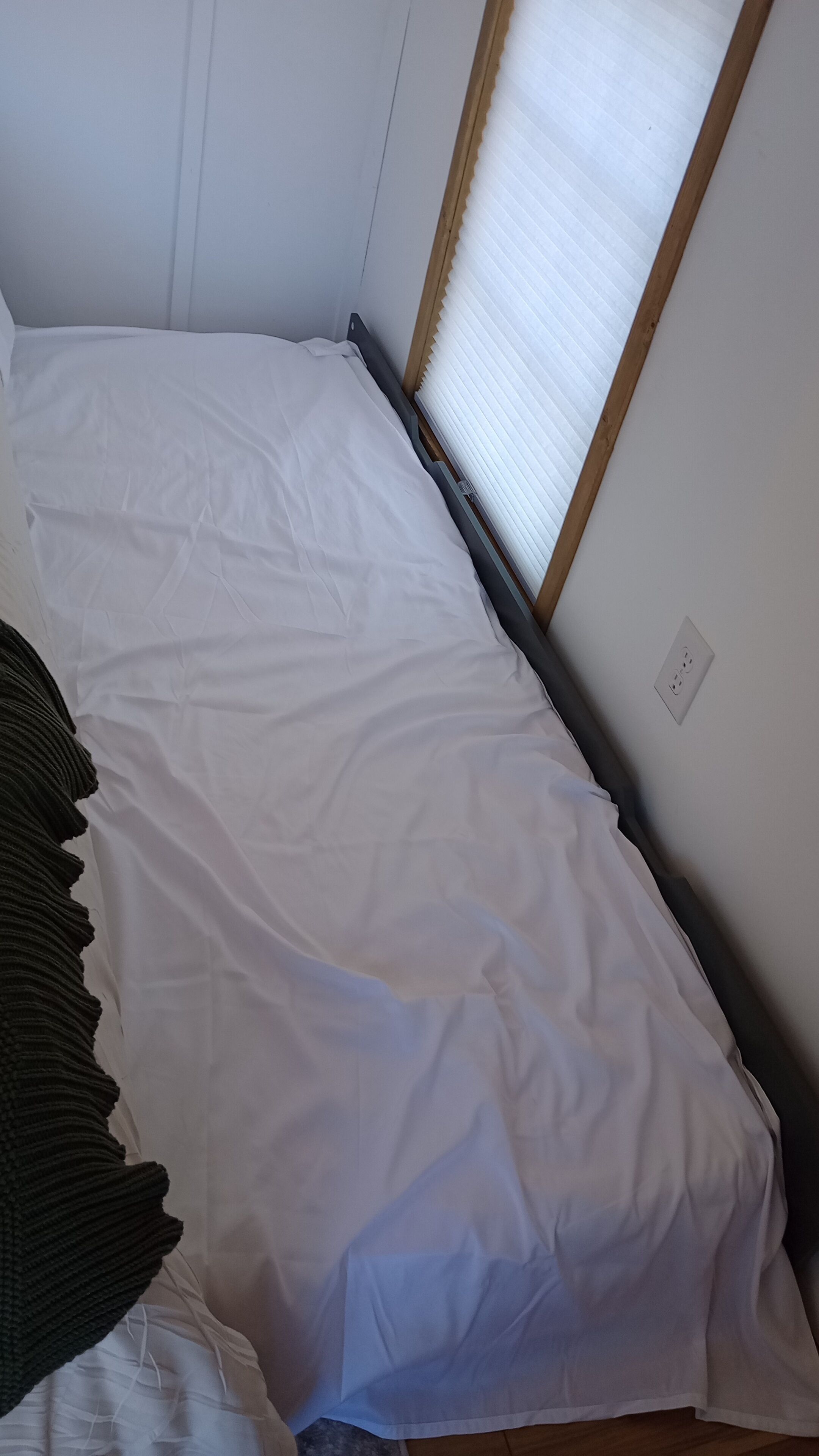 Un lit simple avec une literie blanche, dans une pièce dotée d’une fenêtre et d’une prise de courant au mur.
