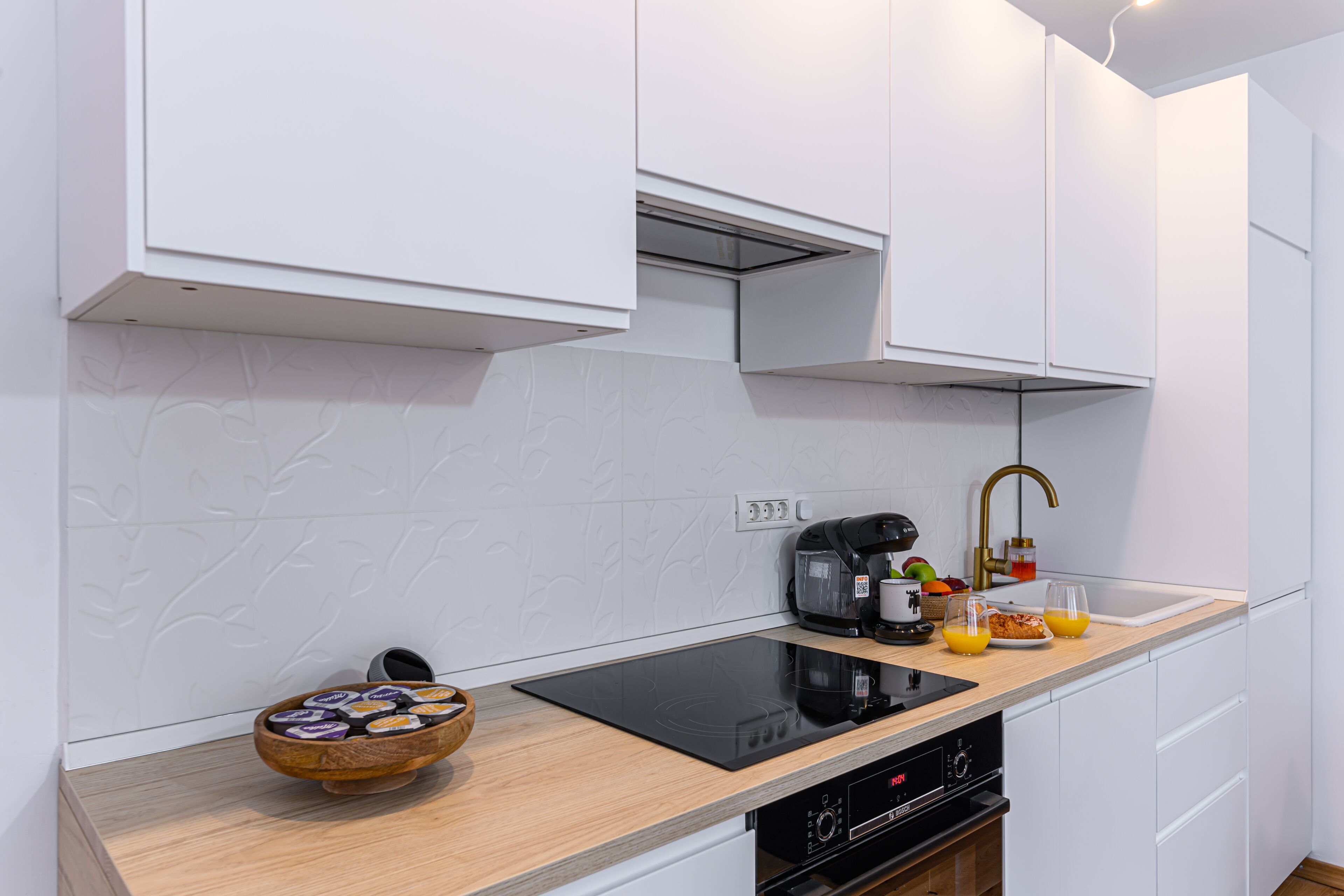 A modern kitchen with white cabinets, a wooden countertop, an induction cooktop, a coffee maker, a sink with a gold faucet, and a bowl of snacks.