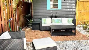 En liten trähydda med ett blått fönster, en flätad soffa med kuddar och en innergård med tegelbeläggning och utemöbler.