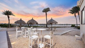 Ein Sitzbereich am Strand mit Reetdachschirmen und weißen Stühlen, mit Blick auf ein ruhiges Meer und einen Sonnenuntergangshimmel.