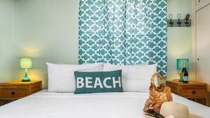 Ein ordentlich bezogenes Bett mit einem Kissen mit der Aufschrift "BEACH", einer Lampe, einer Champagnerflasche, einem Strohhut und einer Strandtasche auf einem Nachttisch aus Holz.