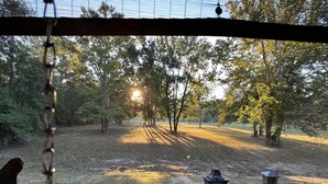 A view from a porch with a swing, overlooking a grassy area with trees and a wood-burning stove.