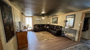 A living room with a black leather sofa, wooden floor, and a large rug. There is a wooden cabinet, a mirror, and a ceiling fan.