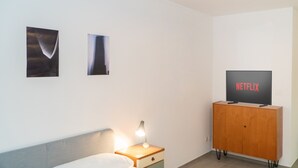 Quarto com cama, mesa de cabeceira com abajur, armário de madeira com uma TV exibindo 'NETFLIX' e duas fotos emolduradas na parede.