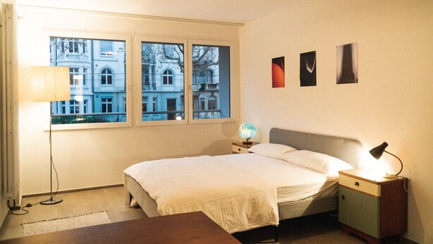 Quarto com cama, abajur, globo terrestre e janela com vista para edifícios.