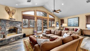 A spacious living room with a stone fireplace, large windows, a ceiling fan, and a cozy seating area. - Steamboat in Style! Dog Friendly, Walk to Resort, Hot Tub, Great Views, Locker (Steamboat Springs)